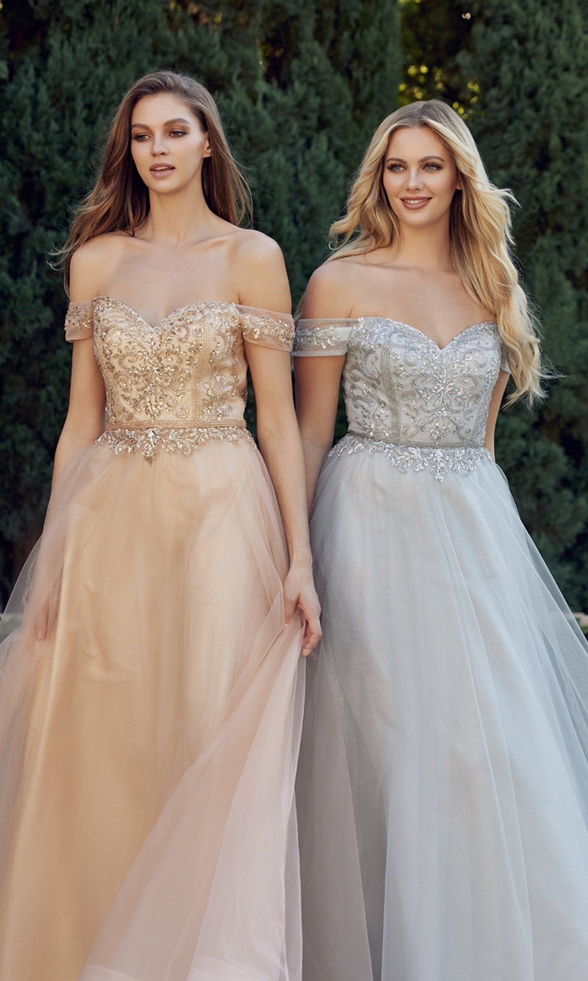 Two women stand outdoors in Long Sweetheart Prom Ball Gown: Juliet 287—floor-length, off-the-shoulder dresses with beaded corset bodices and tulle skirts—one in peach, the other in silver, radiating fairytale elegance.